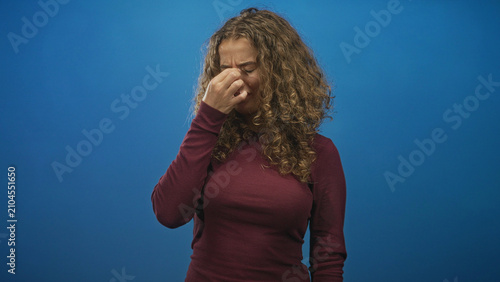 Photos Woman with curly hair wearing a maroon sweater pinches nose with fingers and closes eyes in studio blue backdrop; disgust
