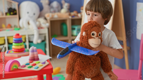 Photography Boy hugging a large stuffed monkey while holding a foam airplane by a colorful toy table in a playroom inside a building; joyful play