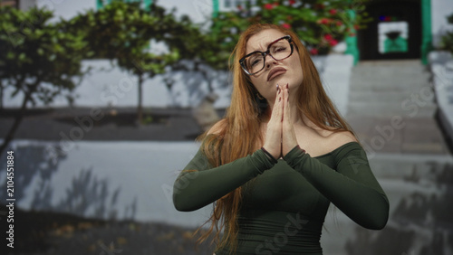 Obraz na plátne Redhead woman presses hands together in pleading prayer at a white building entrance with steps and planters, glasses and green off shoulder top; pleading vulnerability