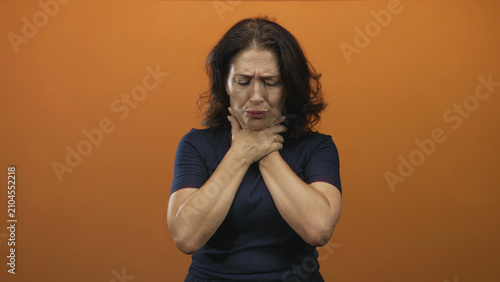 Bild auf Leinwand Middle aged woman clutching neck with both hands in studio orange backdrop, choking gesture and tense expression; panic urgent help