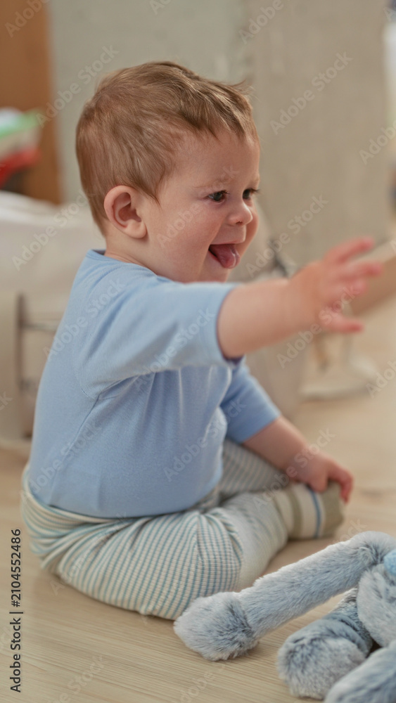 Fototapeta premium Toddler boy reaches with hand and sticks out tongue at plush rabbit while sitting on floor in a building; joy curiosity.