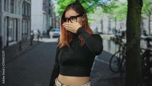 Fotografie Woman covering eyes with hand on street by parked bicycles and tree, wearing sunglasses and black crop top; shyness