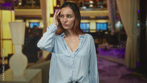 Foto Woman touching head with hand in hotel building lobby, frowning and rubbing temple while wearing a loose light blue shirt; discomfort