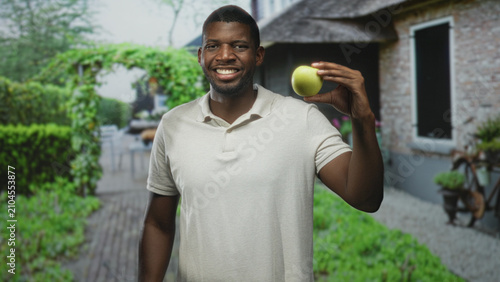 Fotografía Man holds green apple in right hand smiling in front of a residential building courtyard; wellness joy