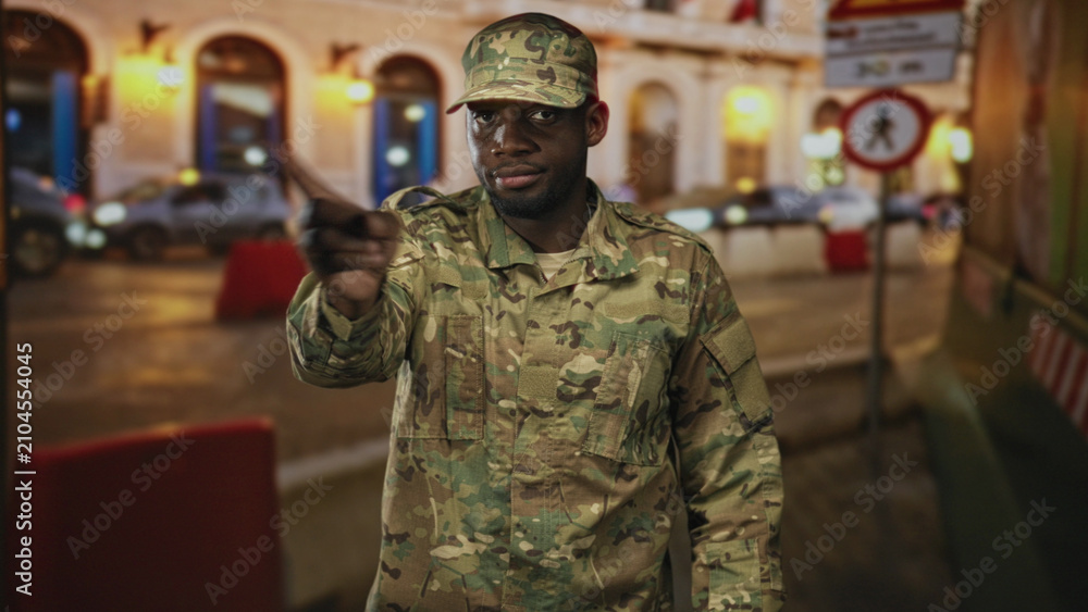 Fototapeta premium Soldier man points finger to camera on city street by lit building arches, visible finger and camo uniform; alert duty vigilance.