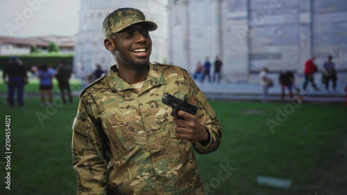 Foto Soldier man in camouflage uniform holding a pistol in one hand, smiling and slightly turning his head while standing on grass before a stone building with blurred tourists; duty