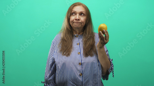 Фотография Woman in striped blouse holds a lemon aloft in her right hand in a green studio, looking unimpressed; resignation reflection