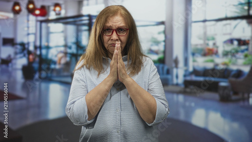 Foto Woman with hands pressed together in prayer gesture wearing red glasses in building lobby, facing camera; pleading concern