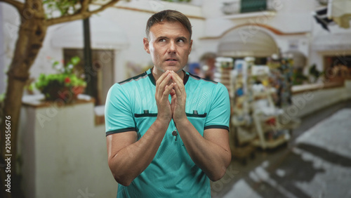 Foto Man referee with whistle wearing turquoise shirt, hands pressed together in pleading gesture on a street market in front of shops and tree; anticipation