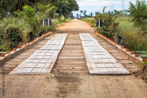 Wooden bridge crossing muddy dirt road through South Pantanal wetlands with lush green vegetation and distant power lines