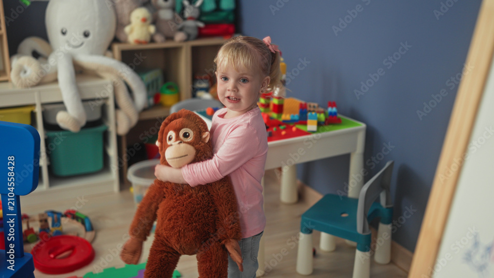 Fototapeta premium Little blonde girl hugging a large stuffed monkey with hands in a studio playroom; playful innocence.
