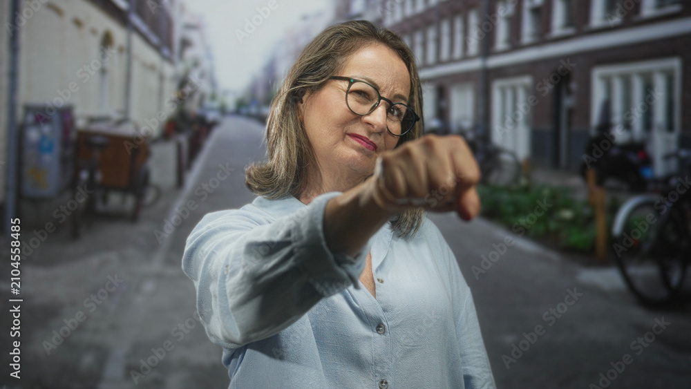 Fototapeta premium Hispanic senior woman with grey hair and glasses extends fist forward on street by bicycles and row houses; confidence empowerment.