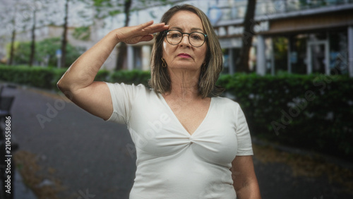 Hispanic senior woman with grey hair wearing a white top salutes with hand to forehead while facing camera on a city street by hedges and benches; pride duty.