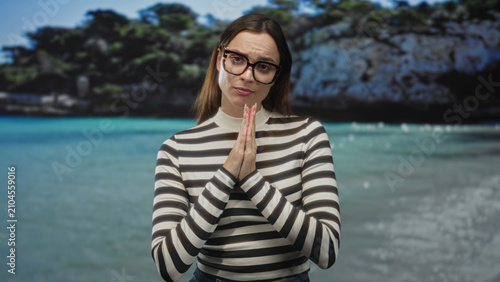 Foto Woman in striped top and glasses with palms pressed together at the beach, pleading expression toward camera; apologetic plea