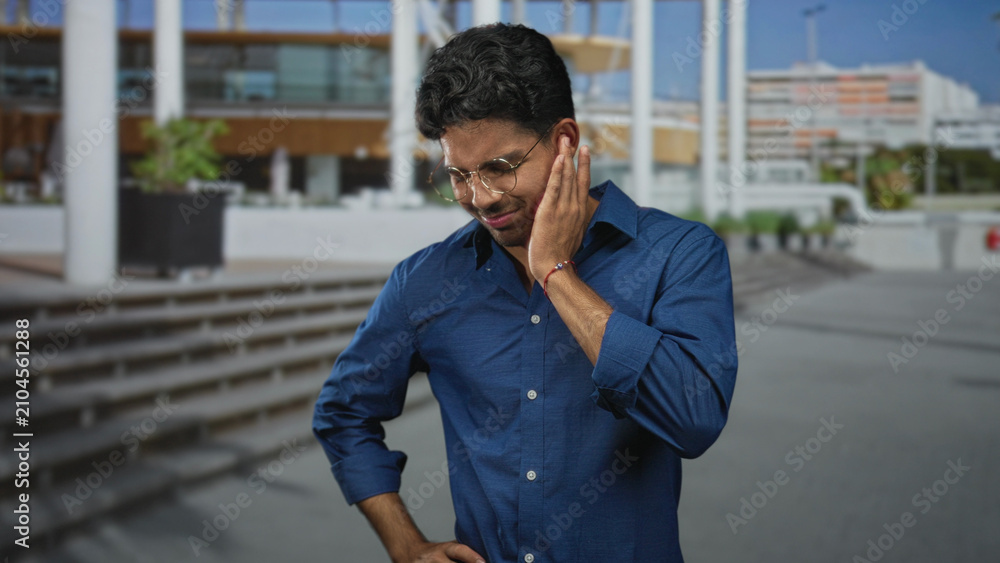 Fototapeta premium Man holding cheek with hand, wearing glasses and blue shirt, wincing near public building steps and plaza entrance; discomfort reflection.