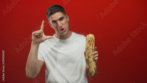 Foto Man holding a baguette and forming an l with his hand in a red studio, squinting and making a mock face; playful teasing