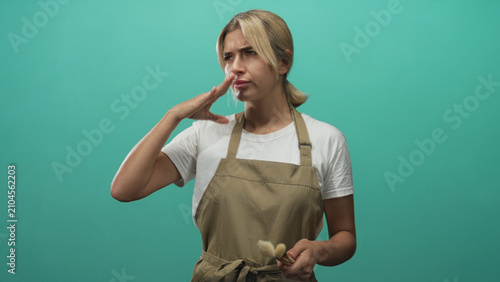 Tableau sur toile Woman makeup artist young blonde wearing apron pinches nose while holding brush in studio with teal backdrop; disgust reaction