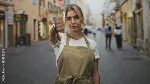 Canvas Print Woman in apron extends thumb up then thumb down toward camera while standing on a busy street with blurred pedestrians and storefronts; ambivalence
