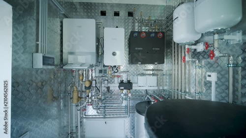 Modern mechanical room interior showing a complex HVAC system with heat pump, boiler, expansion tanks, pipes, and pump groups against a metallic wall.