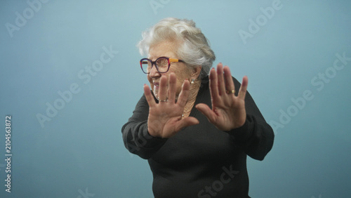 Fotografía Senior woman wearing glasses and rings holds hands palms out in a stop gesture in studio blue backdrop; defensive refusal