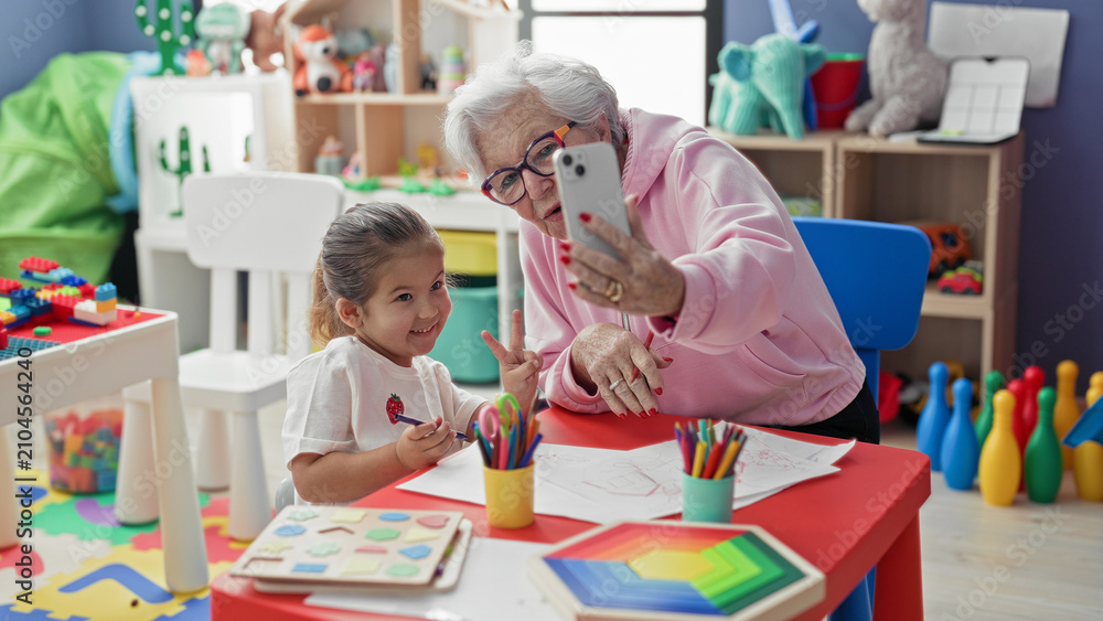 Fototapeta premium Woman holding smartphone taking selfie with little girl at colorful preschool classroom table showing drawings and colored pencils and toys on shelves; generational bonding joy.