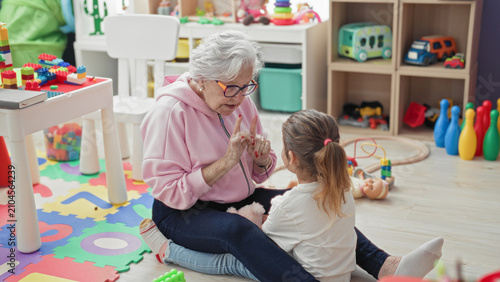 Obraz na płótnie Elderly woman reaches arms to hug little girl on a toy strewn playmat inside a building; affection bonding