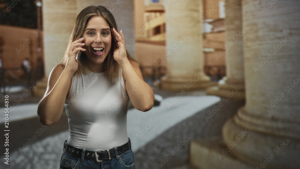 Fototapeta premium Woman speaking on smartphone with hand on hip, wearing white tank top and jeans in historic stone building; cheerful conversation.