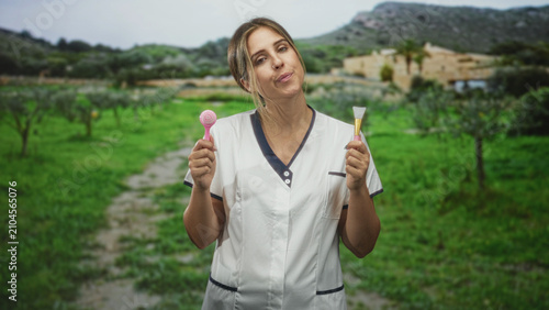 Foto Woman holding a facial cleansing brush and silicone mask applicator while wearing white beautician tunic in front of building on a rural path; serenity care wellness