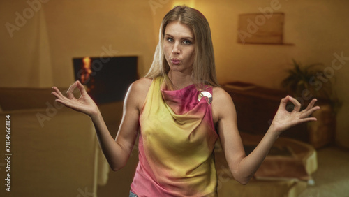 Konstfotografi Woman meditating with hands in mudra beside a sofa in a building, wearing colorful sleeveless top and brooch, eyes closed and relaxed; mindfulness calm