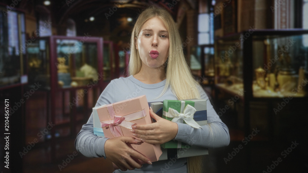 Fototapeta premium Blonde woman smiling and holding wrapped gift boxes with hands across chest inside a display filled building with glass cases and warm lighting; celebration joy.