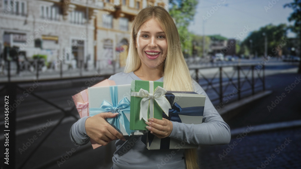 Fototapeta premium Woman holding wrapped gifts with hands across torso on street, smiling broadly while clutching bows; joy celebration.