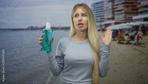 Принти на полотні Woman holding mouthwash bottle, hand raised near nose and lips in a disgusted grimace by a seaside building with umbrellas and sand; disgust caution