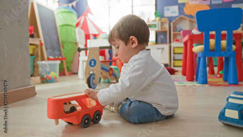 Bild auf Leinwand Boy seated on floor playing with a red toy truck in a bright building playroom; curiosity exploration