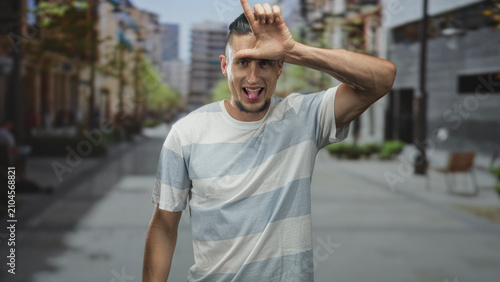 Foto Man sticks out tongue and makes l hand gesture on forehead wearing a striped t shirt on a city street; playful mischief
