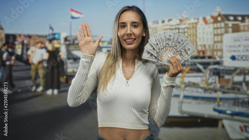 Konstfotografi Woman holding fan of dollar banknotes and waving hand on street dock by boats and canal; financial confidence