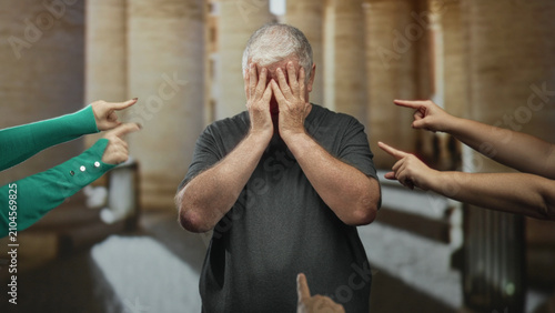 Fotografía Senior man with hand covering face and multiple fingers pointing at him in a building corridor; humiliation