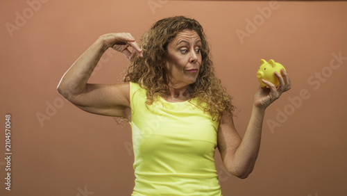 Canvastavla Middle aged hispanic woman holding yellow piggy bank inspects it with pinching finger and raised arm gesture in studio; saving concern