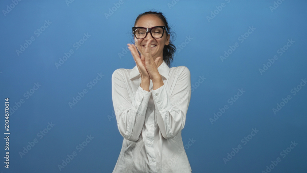 Fototapeta premium Woman clapping hands and smiling in a blue studio, wearing glasses and a white shirt; joy celebration.