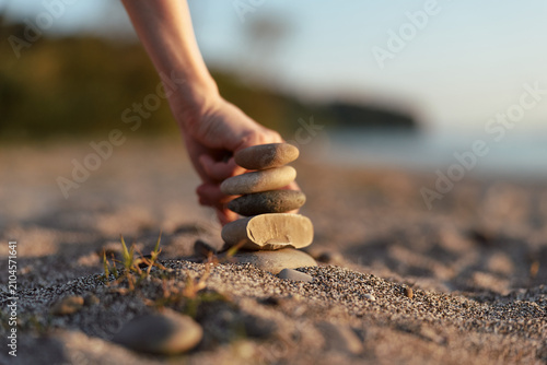 Canvas Print Close up of a hand stacking smooth stones on a sandy beach, warm sunlight glows, creating a sense of balance, calm, and focus
