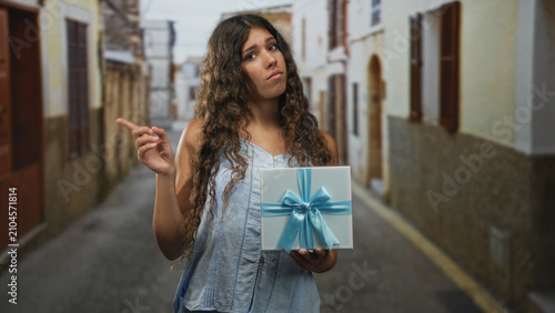 Photos Woman holding gift box with blue ribbon in an old street, sleeveless top and curly hair, offers it with hesitant expression; reluctant offering uncertainty