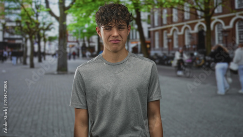 Fotografía Teenage man grimacing face wearing grey tshirt and silver chain on urban street plaza with brick building and trees; discomfort awkwardness