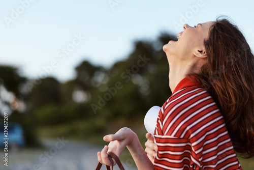 A young woman in a red striped shirt tilts her head back and laughs aloud outdoors, bathed in warm sunlight, conveying carefree mood, spontaneous joy, and summer vitality.