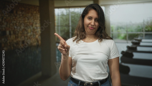 Fényképezés Young hispanic woman points finger at camera by indoor pool in building lobby; casual disapproval gesture