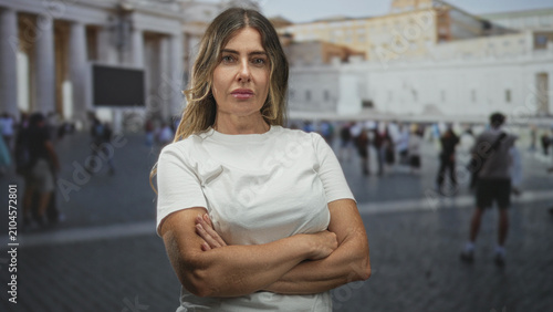 Fotografie Woman stands with crossed arms, showing bare forearms and closed eyes in front of a historic building plaza with crowds and columns; quiet confidence