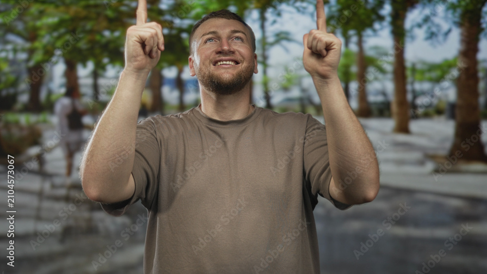 Naklejka premium Man pointing both index fingers up on a tree lined street, smiling and looking upward while wearing a plain brown t shirt; joy celebration.
