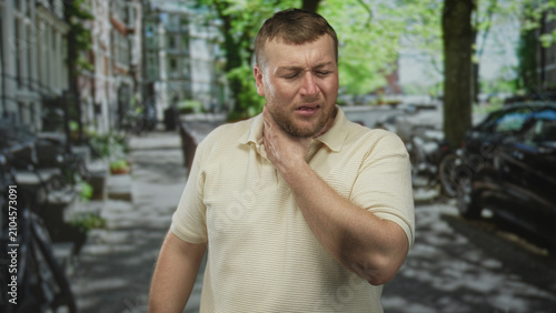 Man clutching his throat with one hand, eyes closed amid bicycles and parked cars on a city street; panic medical emergency.