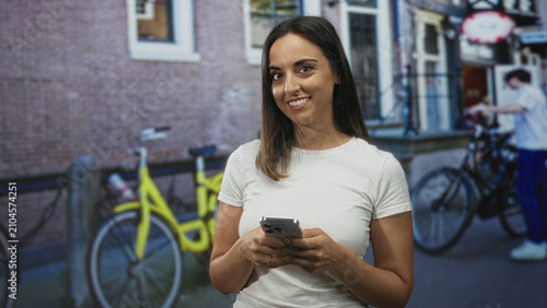 Konstfotografi Woman holding smartphone with both hands, wearing white t shirt, smiling and looking at camera in urban street with bicycles and brick building; contentment connection