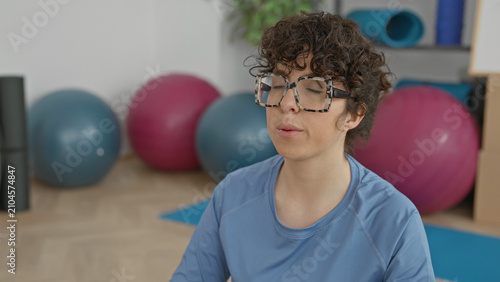 фотографія Woman seated on a blue yoga mat meditating with closed eyes and relaxed face, curly hair and large glasses, exercise balls and foam roller nearby in studio; calm wellness