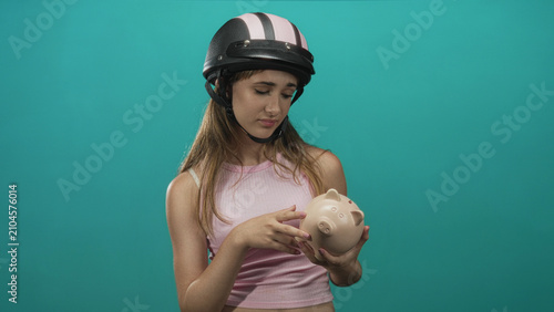 Картината върху платно Young woman wearing helmet holds ceramic piggybank with both hands in turquoise studio while looking down; financial worry