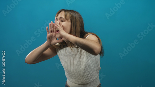 Fotografie Young woman with hands cupped to mouth shouting in a blue studio, leaning forward in a white knitted top; energetic announcement call